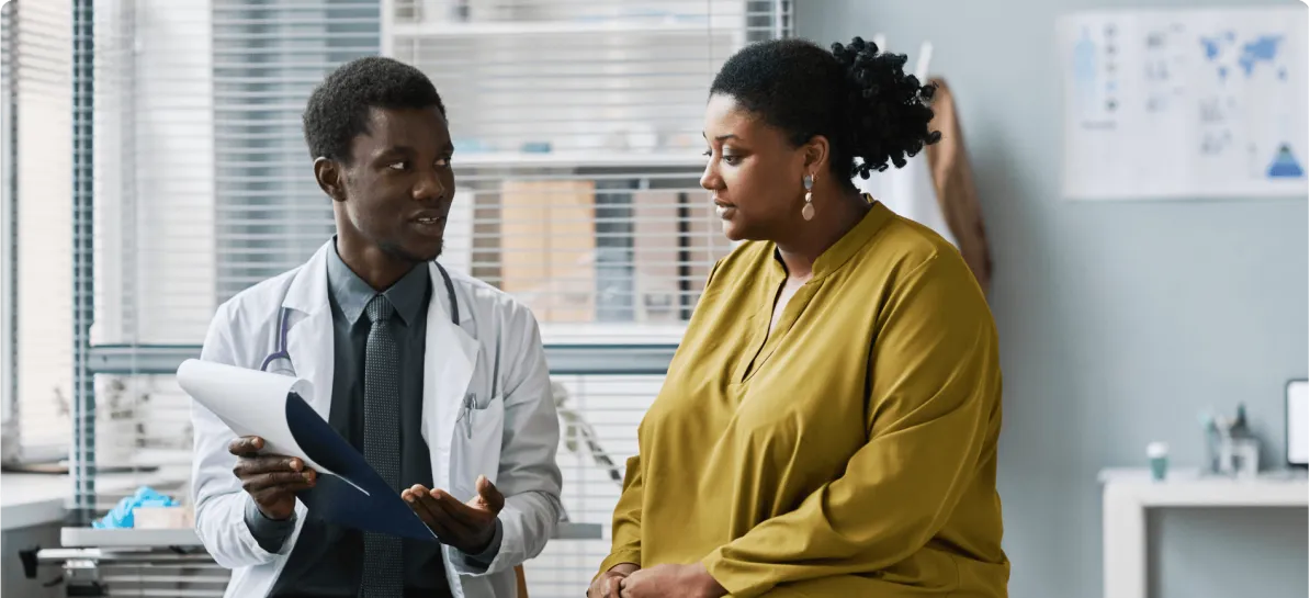 Male doctor in white coat holds up clipboard during a sitting consultation with female patient
